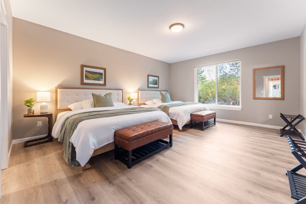 Spacious master bedroom at Bandon's Trail House with two queen beds, ensuite bath access, and forest views through large windows