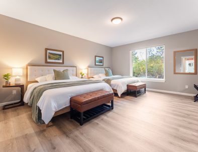 Spacious master bedroom at Bandon's Trail House with two queen beds, ensuite bath access, and forest views through large windows